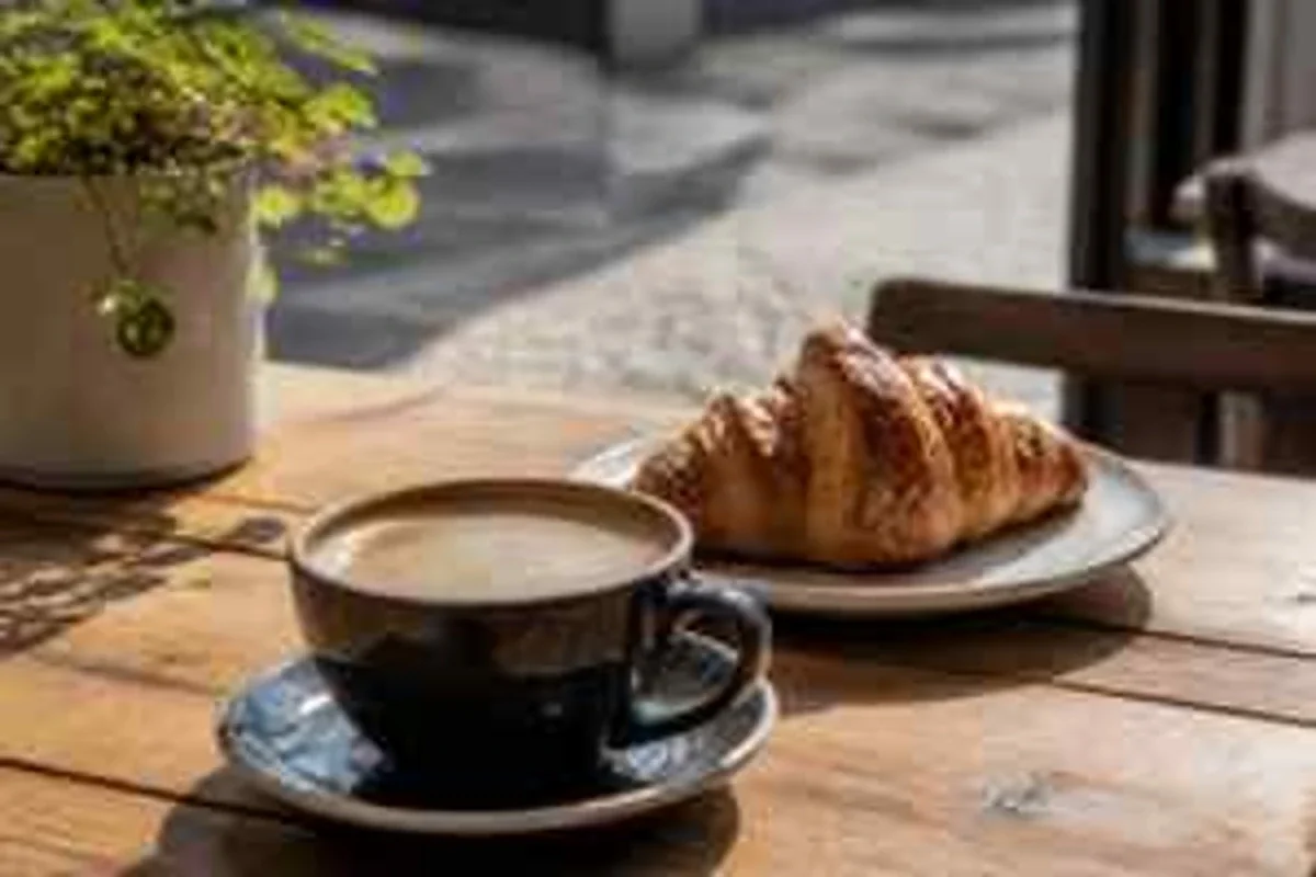 AI-generated photo of a sunlit café table with a coffee and a croissant, artificially degraded, before enhancement